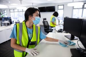 Mixed race woman and colleague wearing hi vis vests, gloves and face masks sanitizing an office using disinfectant. Hygiene in workplace during Coronavirus Covid 19 pandemic. by Wavebreak Media LTD. 